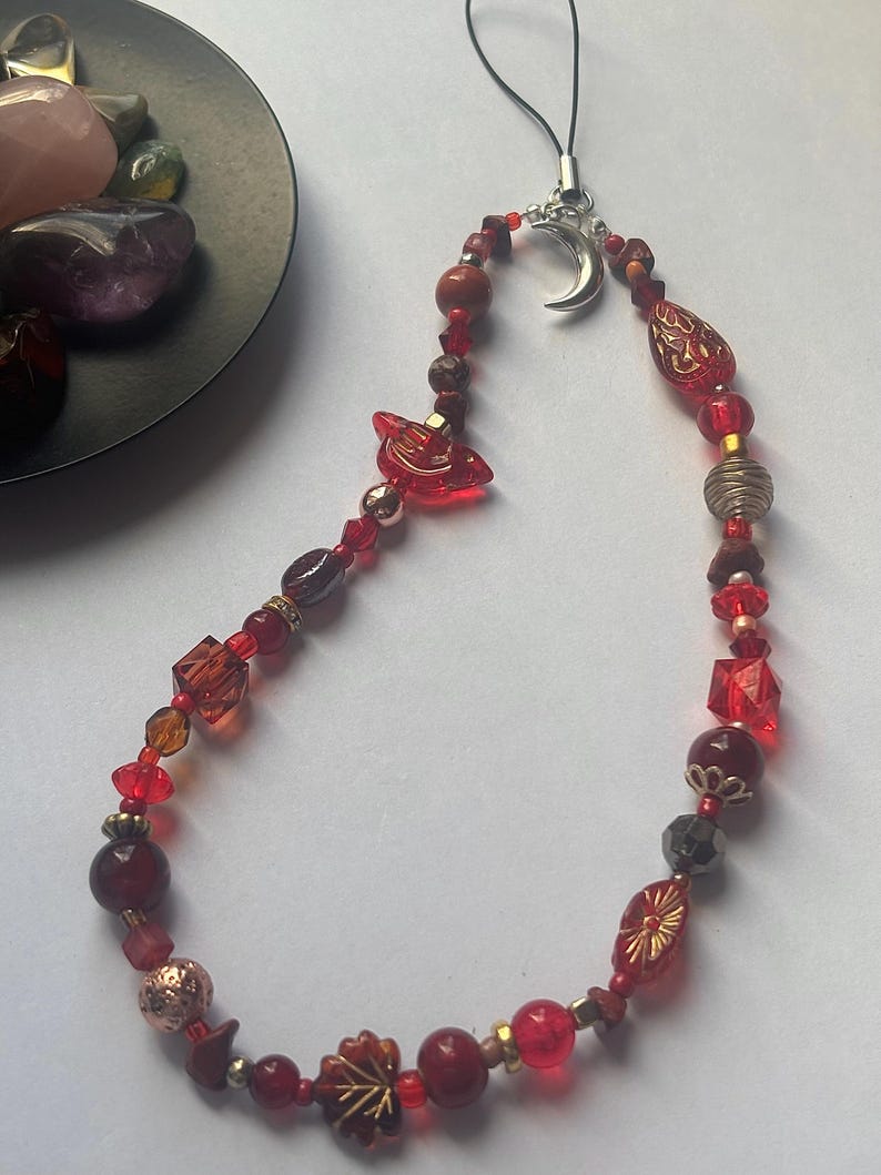 May include: A phone charm featuring a variety of red beads in different shapes, sizes, and textures, including faceted, smooth, and patterned glass beads. A silver-toned crescent moon charm is attached to the top, connected to a black cord loop.