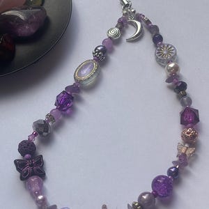 May include: A phone charm featuring a variety of purple, lavender, and clear beads in different shapes and textures. The charm includes silver-toned metal charms shaped like a crescent moon, a spiral, and two butterflies, attached to a black cord.