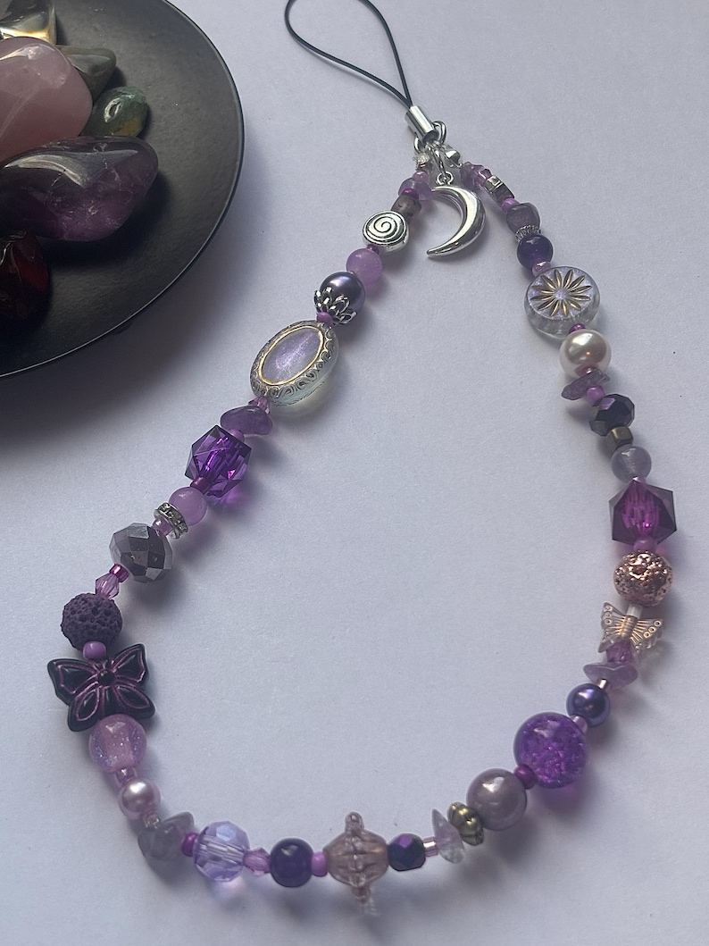 May include: A phone charm featuring a variety of purple, lavender, and clear beads in different shapes and textures. The charm includes silver-toned metal charms shaped like a crescent moon, a spiral, and two butterflies, attached to a black cord.