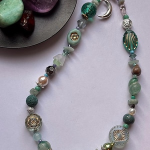 May include: A phone charm featuring a variety of beads in shades of green, teal, and clear glass. The charm includes a silver-toned crescent moon pendant, floral-shaped beads, textured round beads, and small metallic accents strung on a black cord.