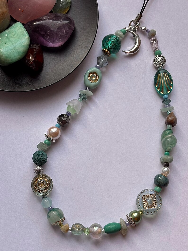 May include: A phone charm featuring a variety of beads in shades of green, teal, and clear glass. The charm includes a silver-toned crescent moon pendant, floral-shaped beads, textured round beads, and small metallic accents strung on a black cord.