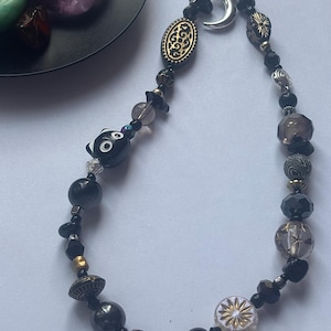 May include: A phone charm featuring a variety of black, grey, and clear beads, including a black cat-shaped bead, a silver crescent moon charm, and an oval gold-toned bead with intricate black patterns, all strung on a thin black cord.