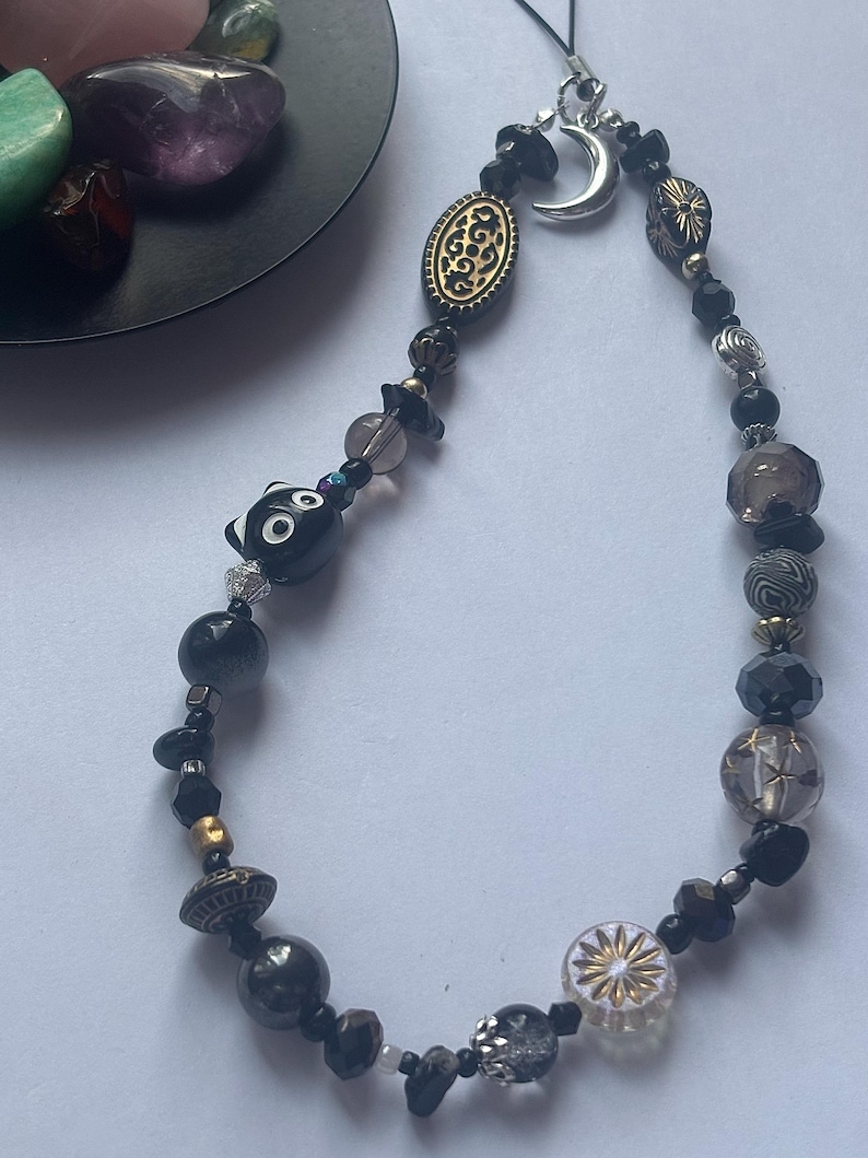 May include: A phone charm featuring a variety of black, grey, and clear beads, including a black cat-shaped bead, a silver crescent moon charm, and an oval gold-toned bead with intricate black patterns, all strung on a thin black cord.