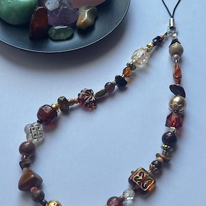 May include: A beaded phone charm with a variety of beads in shades of brown, gold, and clear. The charm includes faceted, round, and square beads, with a black loop for attachment. A small black dish holds several polished gemstones.