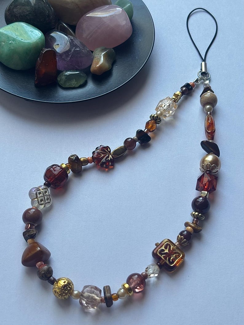 May include: A beaded phone charm with a variety of beads in shades of brown, gold, and clear. The charm includes faceted, round, and square beads, with a black loop for attachment. A small black dish holds several polished gemstones.