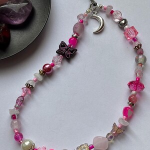 May include: A phone charm featuring a variety of pink, clear, and white beads strung on a black cord. The charm includes a silver crescent moon pendant, a dark butterfly bead, a light pink heart bead, and a small gold-toned butterfly charm.