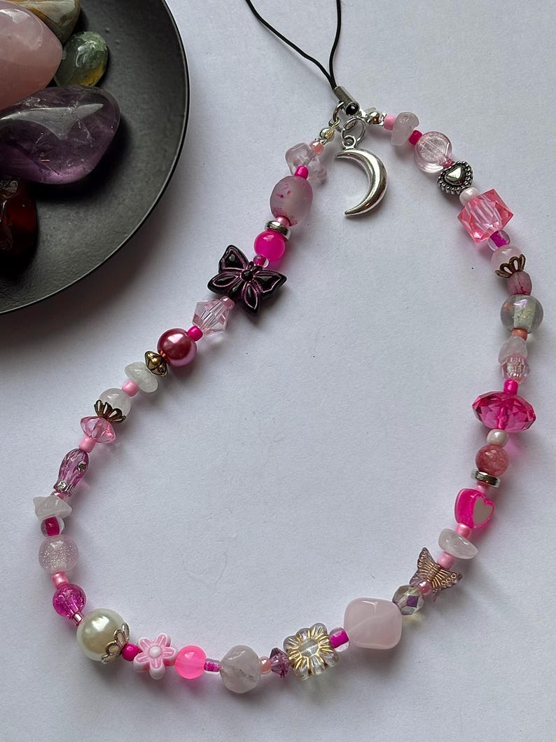 May include: A phone charm featuring a variety of pink, clear, and white beads strung on a black cord. The charm includes a silver crescent moon pendant, a dark butterfly bead, a light pink heart bead, and a small gold-toned butterfly charm.