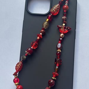 May include: A beaded phone charm with a variety of red beads, including round, square, and decorative shapes. The charm is attached to a black phone case. Gold and bronze accents are interspersed throughout the design.