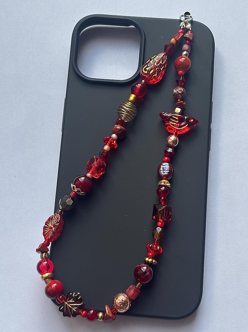 May include: A beaded phone charm with a variety of red beads, including round, square, and decorative shapes. The charm is attached to a black phone case. Gold and bronze accents are interspersed throughout the design.