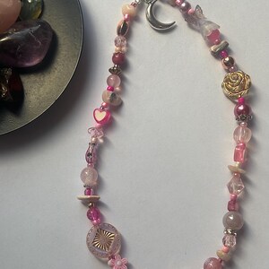 May include: A phone charm strap featuring a variety of pink, clear, and metallic beads. The design includes a silver-toned crescent moon charm, a gold-toned rose bead, a small heart bead, a flower-shaped bead, and a square textured bead.