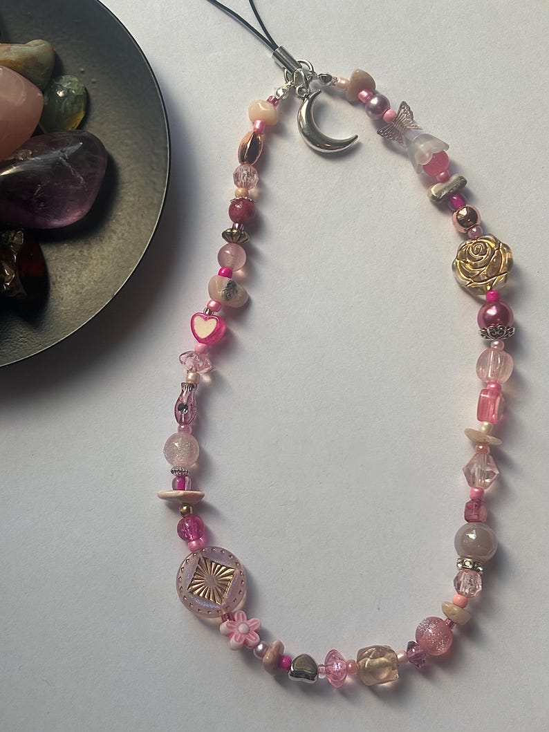 May include: A phone charm strap featuring a variety of pink, clear, and metallic beads. The design includes a silver-toned crescent moon charm, a gold-toned rose bead, a small heart bead, a flower-shaped bead, and a square textured bead.