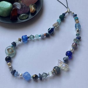 May include: A beaded phone charm featuring a variety of blue, clear, and iridescent beads. The charm includes a mix of round, square, and irregular shaped beads, with a clear bird-shaped bead. A black cord is attached for hanging.