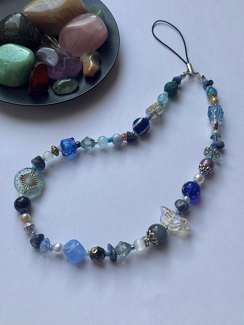 May include: A beaded phone charm featuring a variety of blue, clear, and iridescent beads. The charm includes a mix of round, square, and irregular shaped beads, with a clear bird-shaped bead. A black cord is attached for hanging.