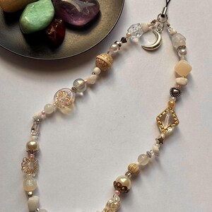 May include: A phone charm featuring a variety of beads in shades of cream, white, gold, and clear. The charm includes a silver crescent moon pendant, a diamond-shaped gold-tone frame with a clear center, and small heart and star accents.