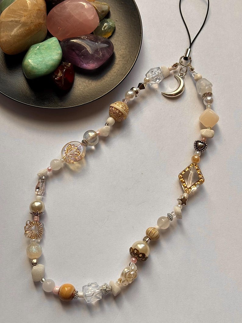May include: A phone charm featuring a variety of beads in shades of cream, white, gold, and clear. The charm includes a silver crescent moon pendant, a diamond-shaped gold-tone frame with a clear center, and small heart and star accents.