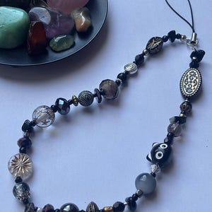 May include: A beaded phone charm featuring a variety of beads in black, silver, and clear. The charm includes a decorative oval bead and a black and white cat bead. A small dish of colorful stones is in the background.