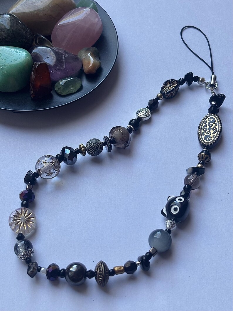 May include: A beaded phone charm featuring a variety of beads in black, silver, and clear. The charm includes a decorative oval bead and a black and white cat bead. A small dish of colorful stones is in the background.