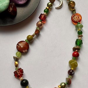 May include: A beaded phone charm featuring a variety of multicolored beads in shades of green, red, brown, and gold. The charm includes a silver-toned crescent moon pendant and a translucent orange bead with a white lotus flower design.