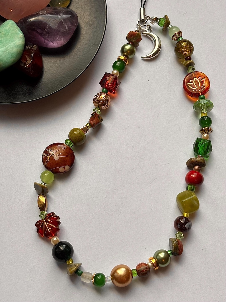 May include: A beaded phone charm featuring a variety of multicolored beads in shades of green, red, brown, and gold. The charm includes a silver-toned crescent moon pendant and a translucent orange bead with a white lotus flower design.