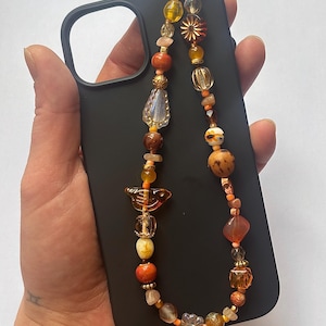 May include: A black phone case with a beaded phone strap. The strap features a variety of orange, yellow, and brown beads in different shapes and sizes. The beads are strung together with a silver-colored wire.