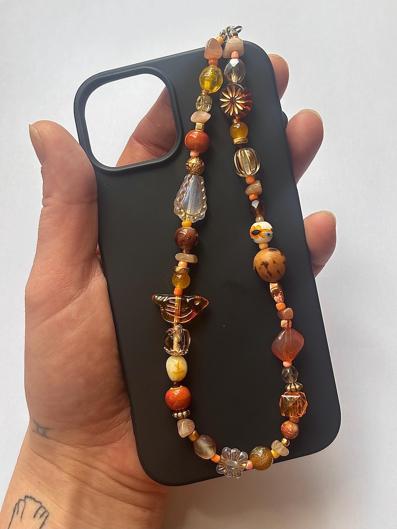 May include: A black phone case with a beaded phone strap. The strap features a variety of orange, yellow, and brown beads in different shapes and sizes. The beads are strung together with a silver-colored wire.