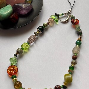 May include: A phone charm featuring a variety of beads in shades of green, brown, and amber, including a silver crescent moon charm, a clear orange bead with a lotus flower design, and a black cord loop for attachment to a mobile device.