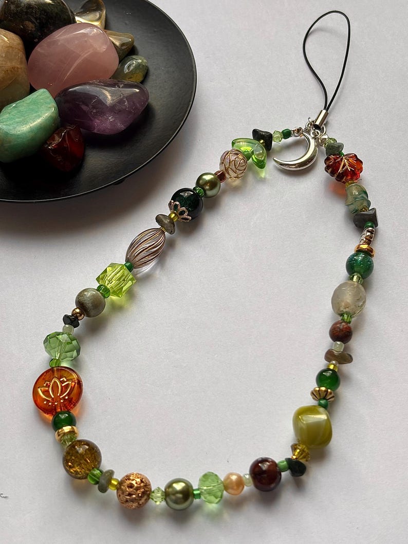 May include: A phone charm featuring a variety of beads in shades of green, brown, and amber, including a silver crescent moon charm, a clear orange bead with a lotus flower design, and a black cord loop for attachment to a mobile device.