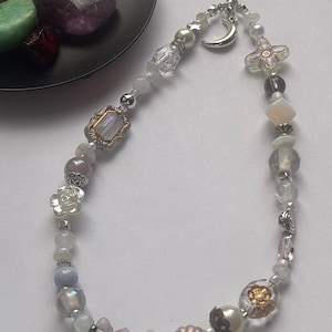 May include: A phone charm featuring a variety of beads including clear, white, and iridescent glass, silver-toned metal accents, a crescent moon charm, and flower-shaped beads. The charm is attached to a black cord loop for easy attachment to devices.