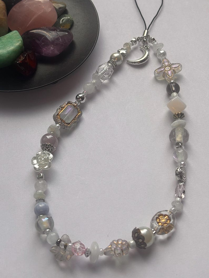 May include: A phone charm featuring a variety of beads including clear, white, and iridescent glass, silver-toned metal accents, a crescent moon charm, and flower-shaped beads. The charm is attached to a black cord loop for easy attachment to devices.