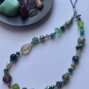 May include: A beaded phone charm featuring a variety of green beads, including faceted crystals, pearls, and decorative floral accents. The charm has a black cord for attachment and includes a mix of opaque and translucent beads.