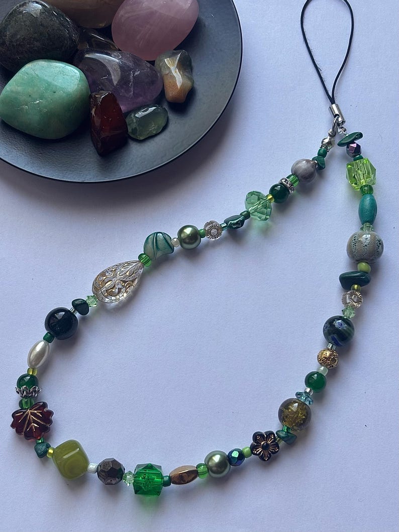 May include: A beaded phone charm featuring a variety of green beads, including faceted crystals, pearls, and decorative floral accents. The charm has a black cord for attachment and includes a mix of opaque and translucent beads.