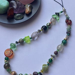May include: A beaded phone charm featuring a variety of green, brown, and clear beads. The charm includes a lotus flower charm and a black cord for attaching to a phone. The beads are various shapes and sizes.