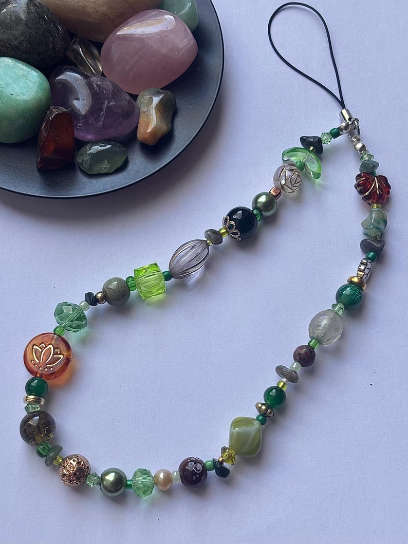 May include: A beaded phone charm featuring a variety of green, brown, and clear beads. The charm includes a lotus flower charm and a black cord for attaching to a phone. The beads are various shapes and sizes.