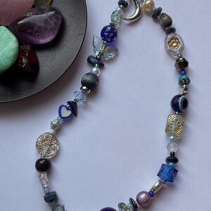 May include: A phone charm featuring a variety of blue, purple, and clear beads, including a silver crescent moon charm, a blue butterfly bead, a heart-shaped bead, and a circular charm with star cutouts, all strung on a black cord loop.