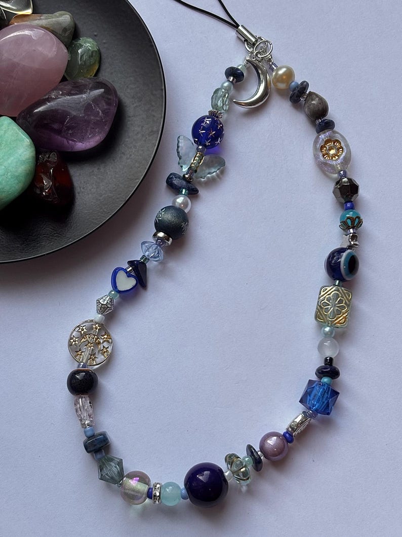 May include: A phone charm featuring a variety of blue, purple, and clear beads, including a silver crescent moon charm, a blue butterfly bead, a heart-shaped bead, and a circular charm with star cutouts, all strung on a black cord loop.
