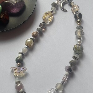 May include: A phone charm strap featuring a variety of beads in shades of gray, white, and clear. The charm includes a silver-toned crescent moon pendant, a clear bird-shaped bead, and several faceted, round, and textured beads strung on a black cord.