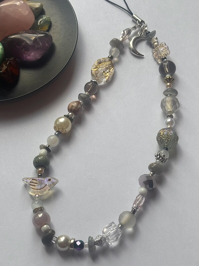 May include: A phone charm strap featuring a variety of beads in shades of gray, white, and clear. The charm includes a silver-toned crescent moon pendant, a clear bird-shaped bead, and several faceted, round, and textured beads strung on a black cord.