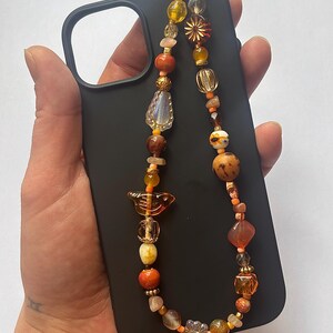 May include: A black smartphone case featuring a decorative beaded strap. The strap is composed of various beads in shades of orange, yellow, brown, and clear, including a bird-shaped charm, flower-shaped beads, and metallic gold accents.