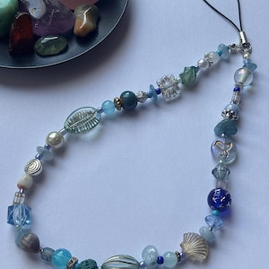 May include: A beaded phone charm featuring a variety of blue and clear beads, including a seashell, flower, and star-shaped beads. The charm has a black loop for attaching to a phone or other device. A small dish of colorful stones is in the background.