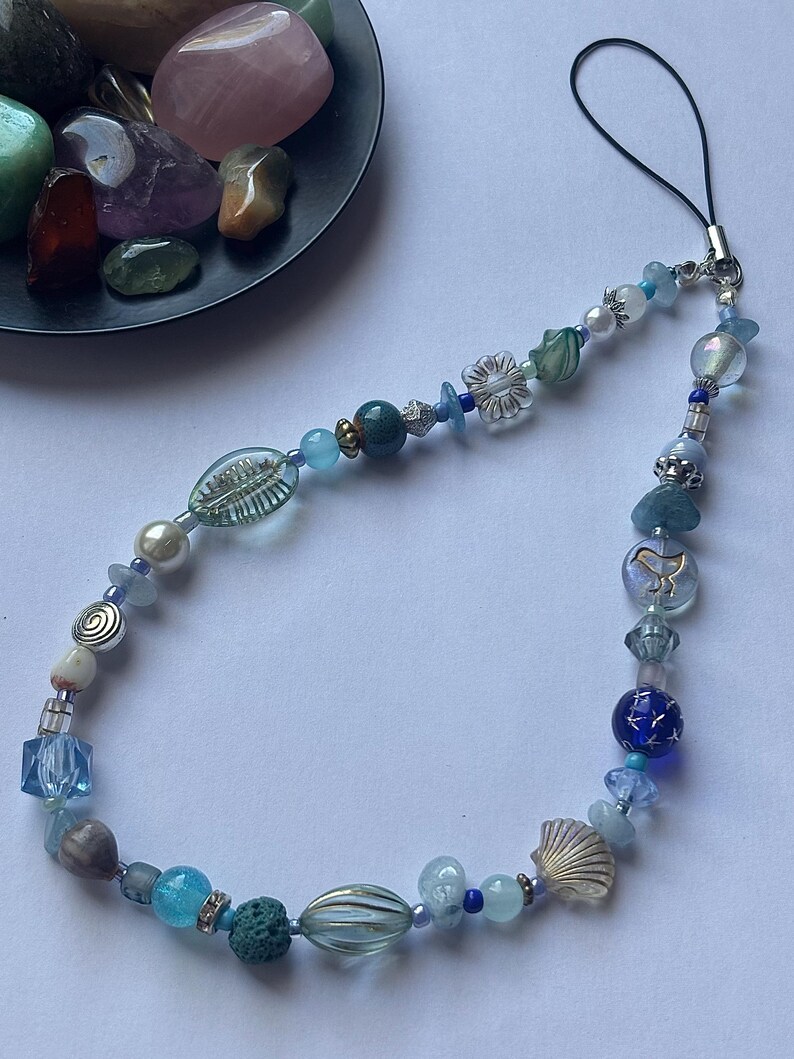 May include: A beaded phone charm featuring a variety of blue and clear beads, including a seashell, flower, and star-shaped beads. The charm has a black loop for attaching to a phone or other device. A small dish of colorful stones is in the background.
