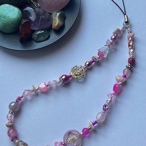 May include: A beaded phone charm featuring a variety of pink beads, including hearts, flowers, and geometric shapes. A gold-colored rose charm is a focal point. The charm has a black cord for attachment.