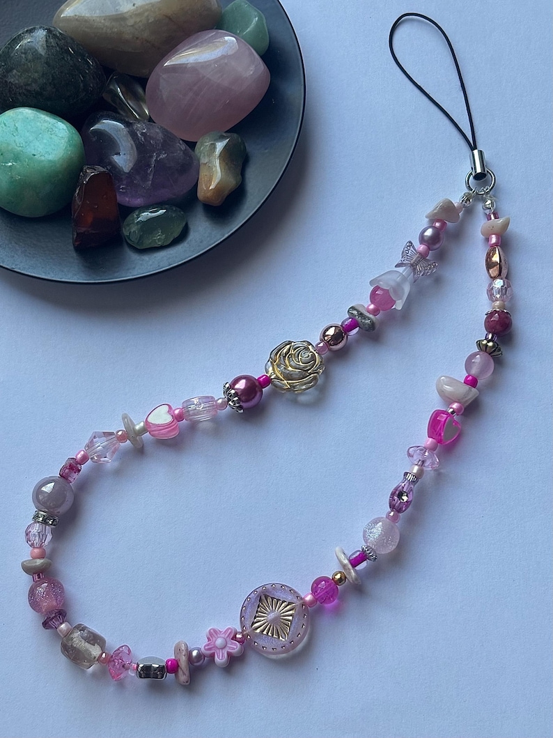 May include: A beaded phone charm featuring a variety of pink beads, including hearts, flowers, and geometric shapes. A gold-colored rose charm is a focal point. The charm has a black cord for attachment.