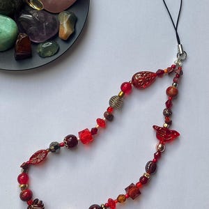 May include: A beaded phone charm featuring a variety of red, gold, and brown beads. The charm includes faceted beads, round beads, and decorative elements. A black loop is attached for easy phone attachment. A dark gray plate with colorful stones is in the background.
