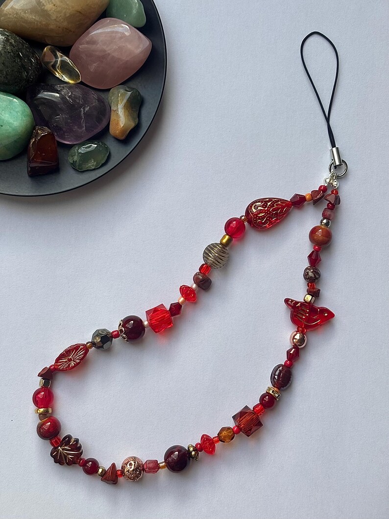 May include: A beaded phone charm featuring a variety of red, gold, and brown beads. The charm includes faceted beads, round beads, and decorative elements. A black loop is attached for easy phone attachment. A dark gray plate with colorful stones is in the background.
