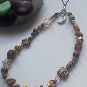 May include: A phone charm featuring a variety of beads in shades of brown, amber, and clear glass, accented with metallic spacers and a silver-toned crescent moon charm. The beads include faceted, smooth, and leaf-shaped designs on a black cord.