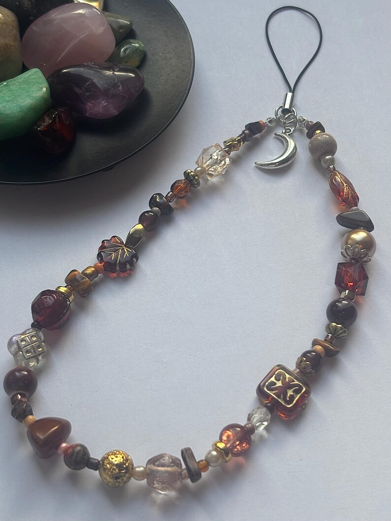 May include: A phone charm featuring a variety of beads in shades of brown, amber, and clear glass, accented with metallic spacers and a silver-toned crescent moon charm. The beads include faceted, smooth, and leaf-shaped designs on a black cord.