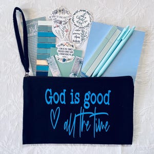 May include: A black zippered pouch with the text "God is good all the time" in blue script. The pouch contains blue pens, stickers, bookmarks, and a small notebook. A black wrist strap is attached to the pouch.