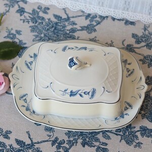 Può includere: Un burriera in ceramica bianca con base rettangolare e coperchio abbinato caratterizzato da un motivo floreale blu e un pomello a forma di fiore scolpito. Il piatto presenta un design a reticolo strutturato sugli angoli e una finitura liscia.