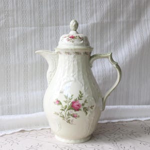 May include: Cream-colored porcelain coffee pot with a lid, handle, and spout. The pot features embossed vertical lines and a floral design with pink roses and green leaves. The handle and rim have a subtle green accent.
