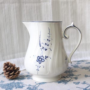 Puede incluir: Jarra de cerámica blanca con diseño floral azul. La jarra tiene un asa curva y un pico. El diseño floral incluye un grupo de flores y tallos con hojas. La jarra está sobre un mantel con estampado azul y blanco.